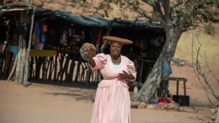 Woman in a pale pink dress holding up a colorful handmade item in front of a rustic roadside stall.