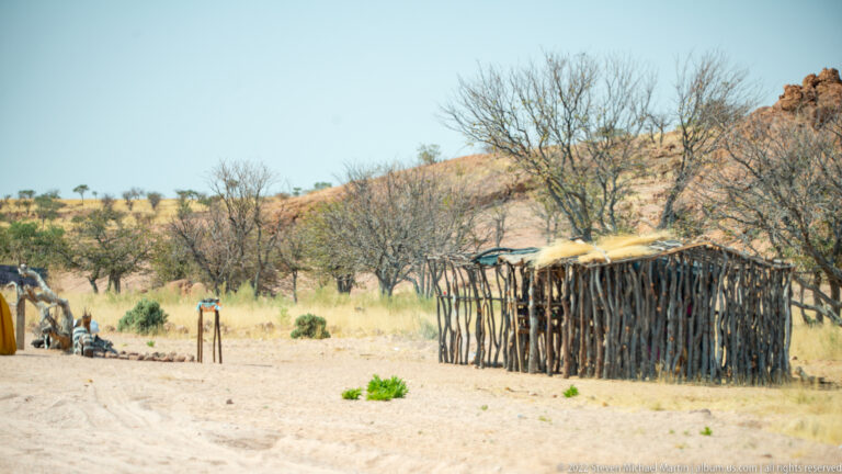 Rectangular branch-built roadside shelter on sandy ground among dry grass and leafless trees.