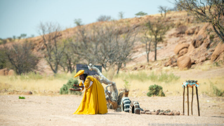 Woman in a bright yellow dress bending beside a roadside display with painted tires in a dry rocky landscape.