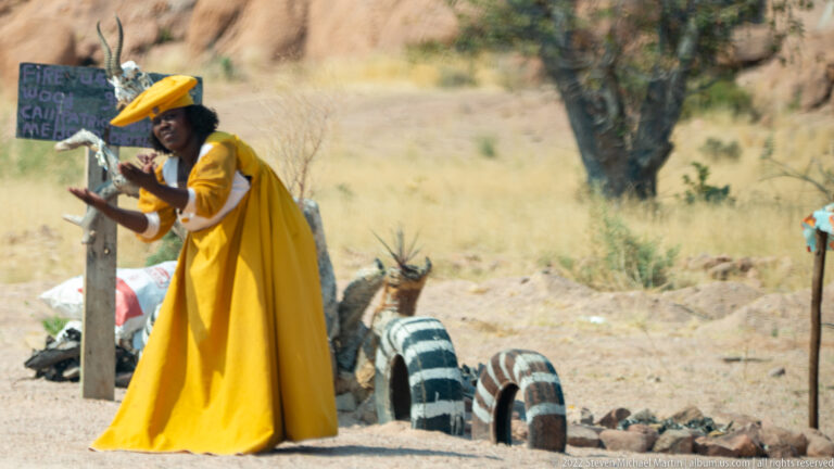 Woman in a bright yellow dress gesturing beside a simple roadside display, with painted tires and goods behind her.