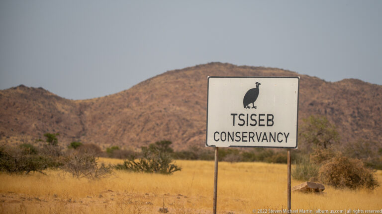 White roadside sign reading TSISEB CONSERVANCY with a black bird emblem, backed by golden grass and a rocky mountain.