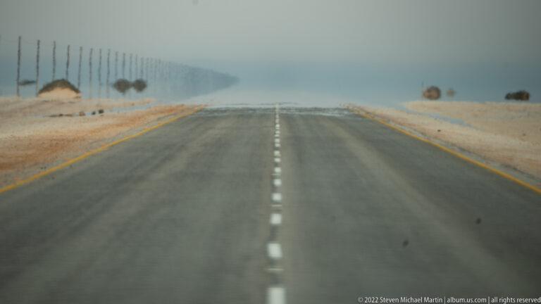 Telephoto view down a straight paved desert highway, with distant poles and roadside features distorted by haze.