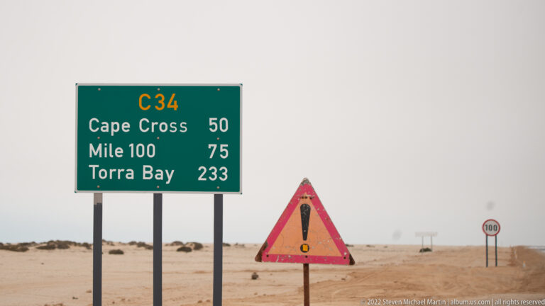 Green C34 distance sign listing Cape Cross 50, Mile 100 75, and Torra Bay 233 beside a desert road.