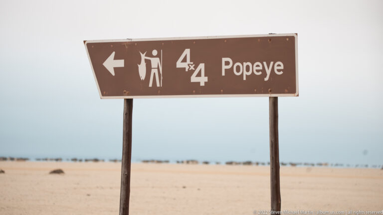 Brown sign pointing left to Popeye, with 4x4 and hiker symbols, against a pale coastal desert background.