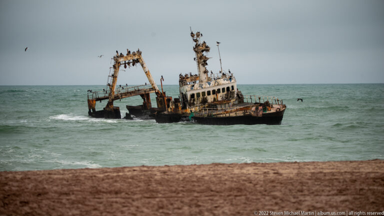 Rusting shipwreck listing in shallow Atlantic water near shore, with many seabirds perched on its mast and superstructure.
