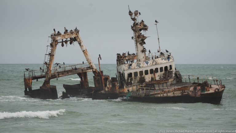 Close view of a rusted shipwreck with tilted wheelhouse and open stern framework in shallow surf, crowded with seabirds.