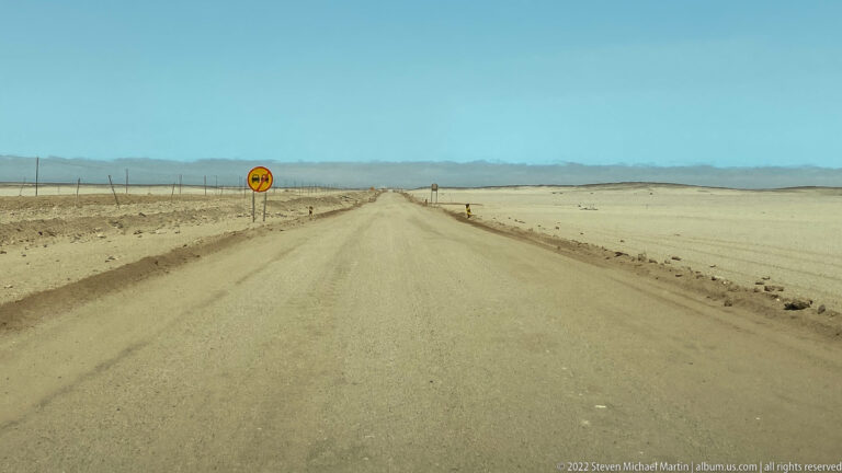 Straight pale gravel road crossing a barren plain toward distant mountains, with roadside signs and a fence line.