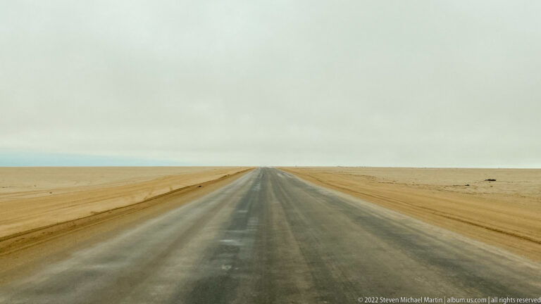 Straight asphalt road vanishing across flat ocher desert beneath a low pale gray sky.