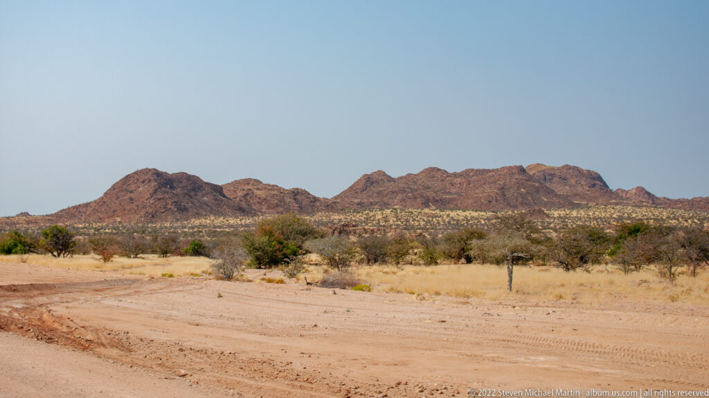 Reddish rocky mountain ridge beyond a flat scrub-covered plain and sandy roadside foreground.