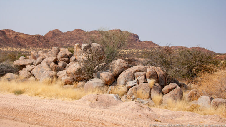 Cluster of large rounded pale boulders beside a gravel road, with dry grass, shrubs, and reddish hills beyond.