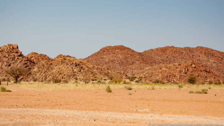 Reddish rocky hills beyond a broad sandy plain dotted with sparse grasses, shrubs, and small trees.