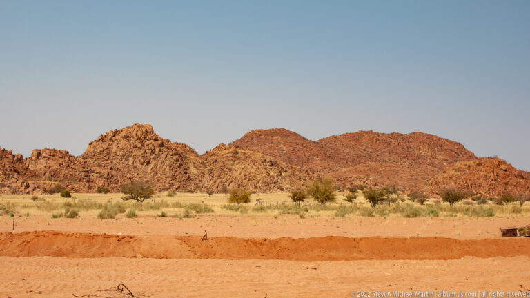 Low reddish rocky ridges beyond dry grassland, with a red soil road cut across the foreground.