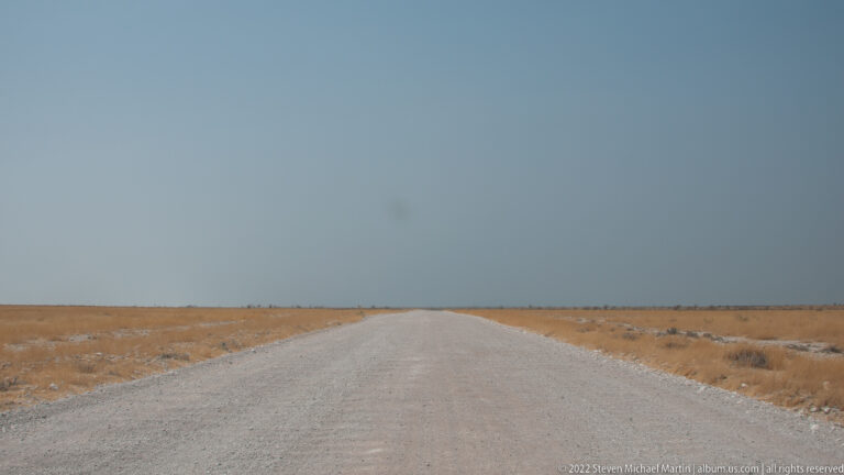 Long straight pale road through flat golden terrain beneath a vast blue-gray sky and low horizon.