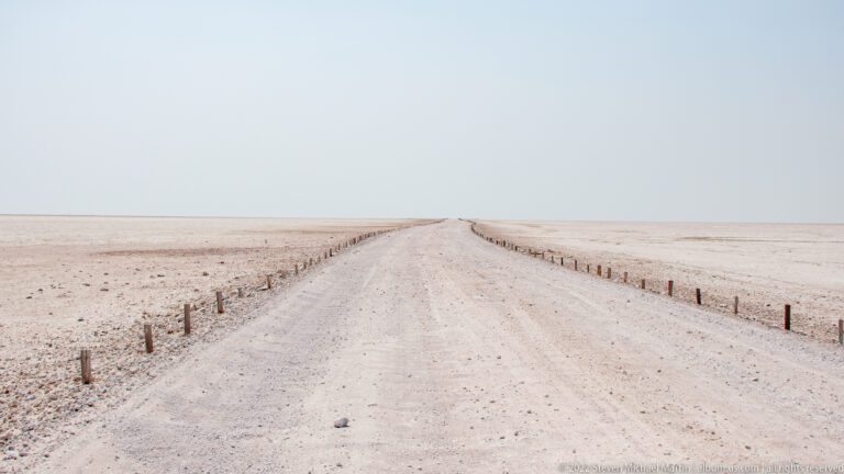 Raised gravel road lined with short posts crossing the pale salt pan toward a hazy horizon.