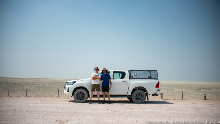 Two travelers stand beside a white pickup truck on the pale Etosha Pan beneath a cloudless blue sky.