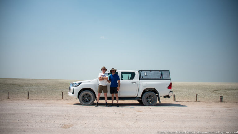 Two travelers and a white pickup appear small against a broad blue sky and the flat pale surface of Etosha Pan.