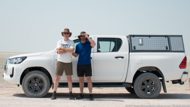 Two travelers in hats and sunglasses stand in front of a white pickup with a metal canopy on Etosha Pan.