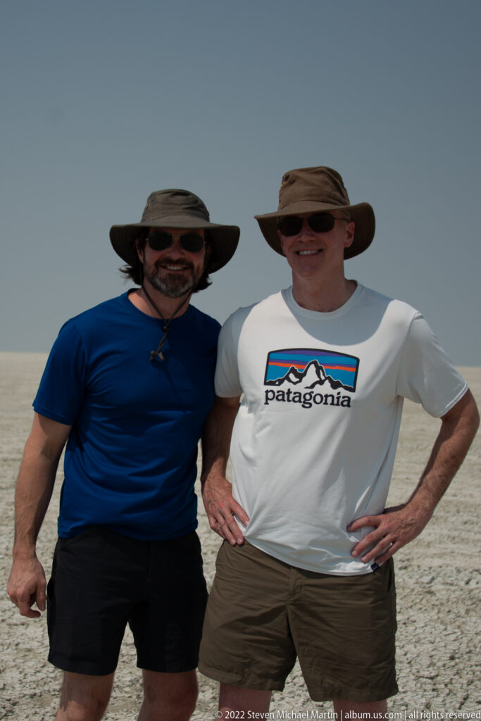 Two travelers in broad-brimmed hats and sunglasses pose together against the flat pale horizon of Etosha Pan.