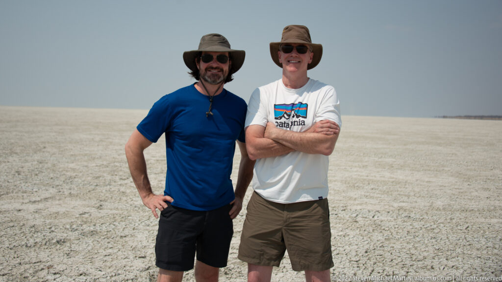 Two travelers stand full length on the rough pale salt surface of Etosha Pan, wearing hats, sunglasses, shirts, and shorts.