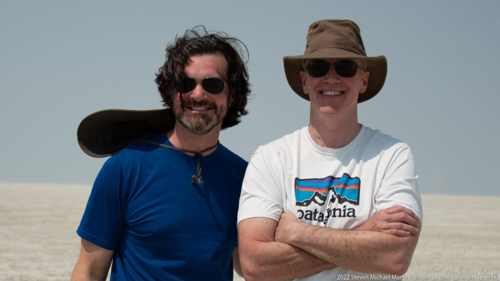 Two smiling travelers in sunglasses are framed waist-up on Etosha Pan, with one hat brim blown upward by wind.