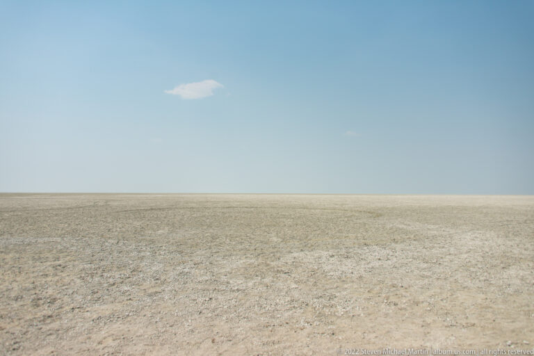 Pale textured salt pan extends to a flat horizon beneath blue sky with a single small white cloud.