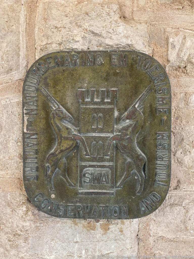Weathered metal conservation plaque on stone wall with wildlife emblems, a central fort motif, “SWA,” and “CONSERVATION” lettering.