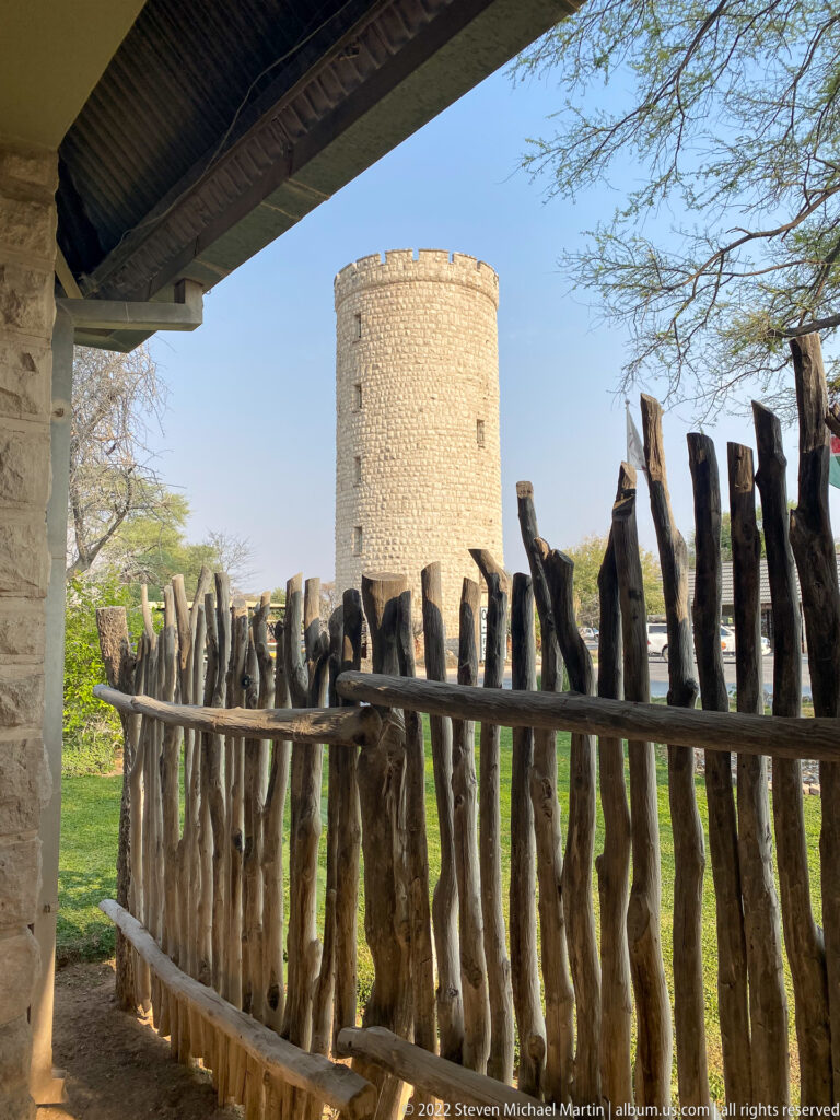 Round pale-stone tower rises behind a rough vertical-log fence, viewed from beneath a shaded roof at an Etosha visitor area.