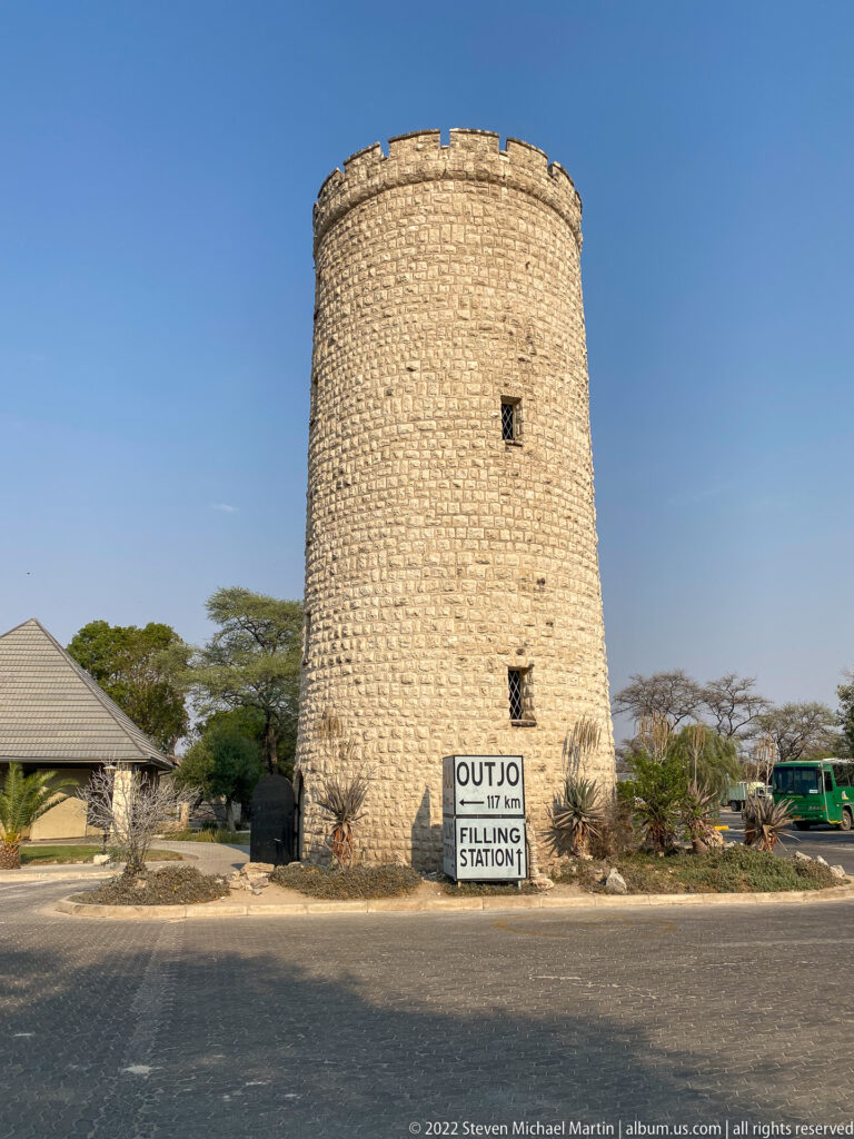 Tall cylindrical stone tower with small windows and a crenellated top stands beside a sign pointing to Outjo and a filling station.