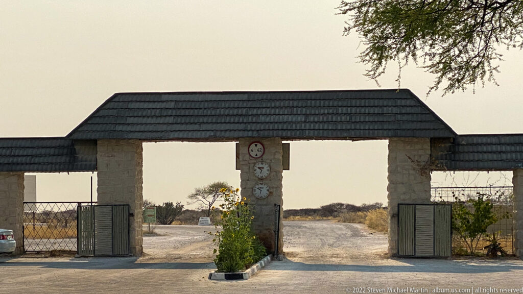 Roofed stone gateway with two open vehicle passages and metal gates leads toward a pale road through dry woodland.