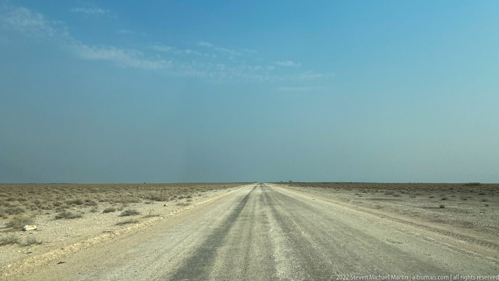 Straight gravel road narrows toward the horizon through low scrub and pale stony terrain beneath a blue sky.