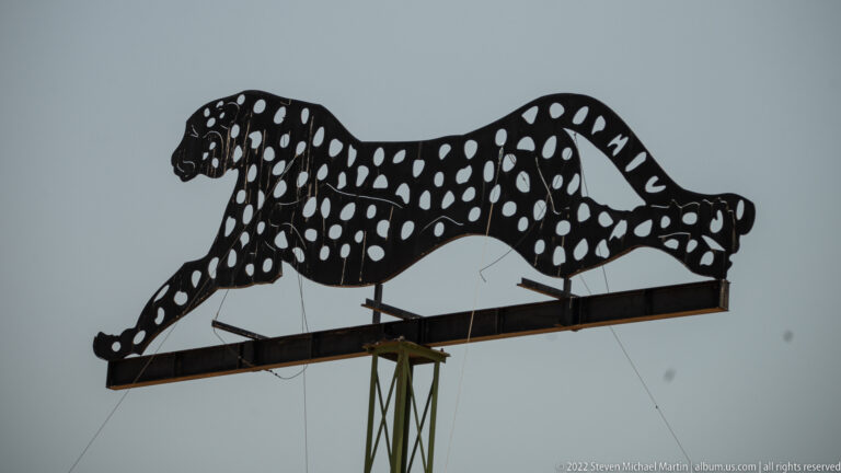 Large black cut-metal running cheetah silhouette with white spots mounted high on a roadside support.