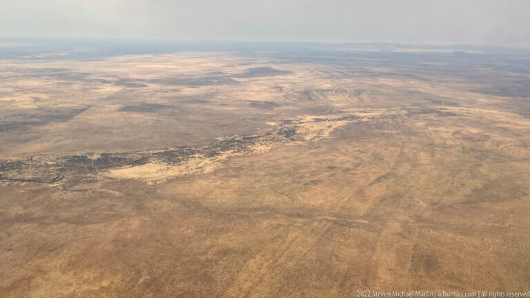 Aerial view across vast tan semi-arid plains with sparse vegetation, pale patches and a hazy distant horizon.