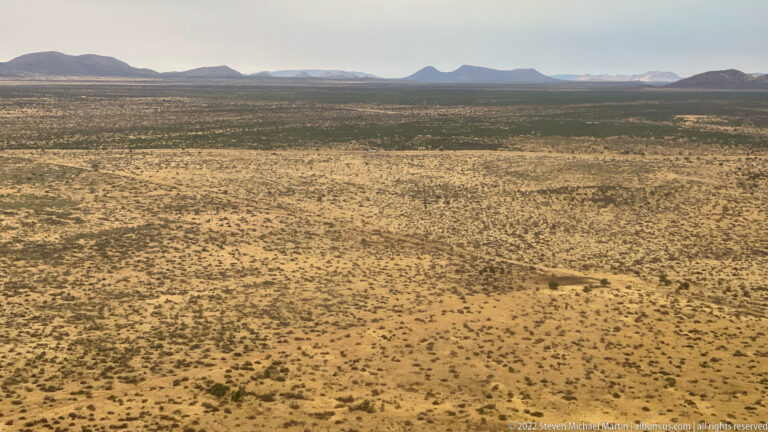 Aerial view over pale shrub-dotted savanna toward isolated dark hills and ridges on the horizon.
