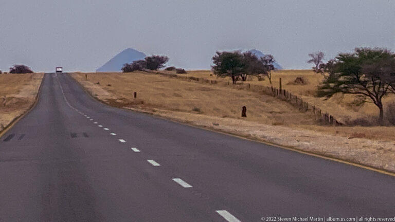Straight paved highway through dry golden savanna with roadside fences, scattered trees and a pointed hill ahead.