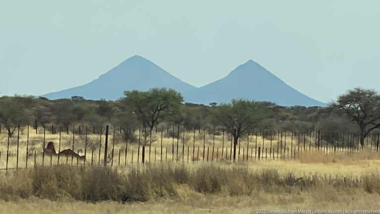 Two conical hills beyond dry grassland, thorn trees and a roadside fence in northern Namibia.