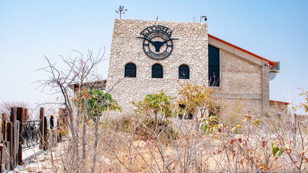 Stone Etosha Trading Post building with buffalo emblem, arched windows, red roof trim and dry thorn scrub.