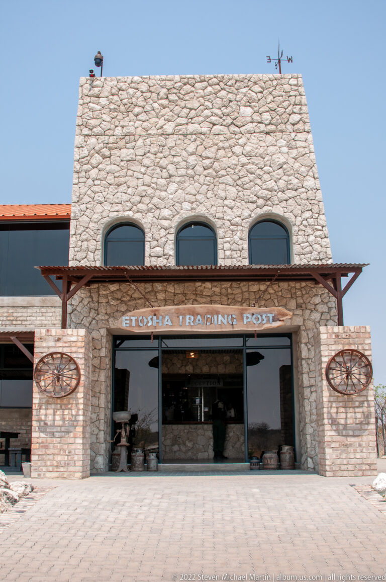 Front entrance of Etosha Trading Post with stone facade, timber canopy, glass doors and wagon-wheel decorations.