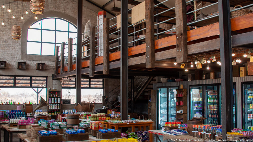 High-ceilinged trading post shop with timber framing, stone walls, drink refrigerators and tables of packaged goods.