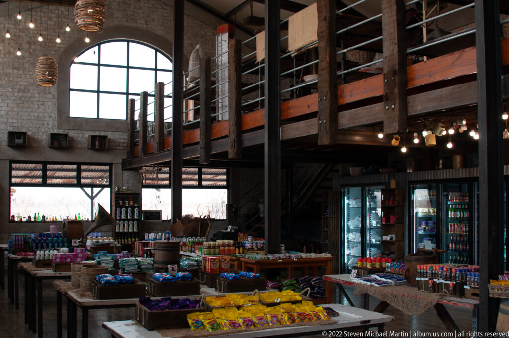 Tables of snacks and provisions inside a timber-and-stone trading post, with illuminated drink refrigerators behind.