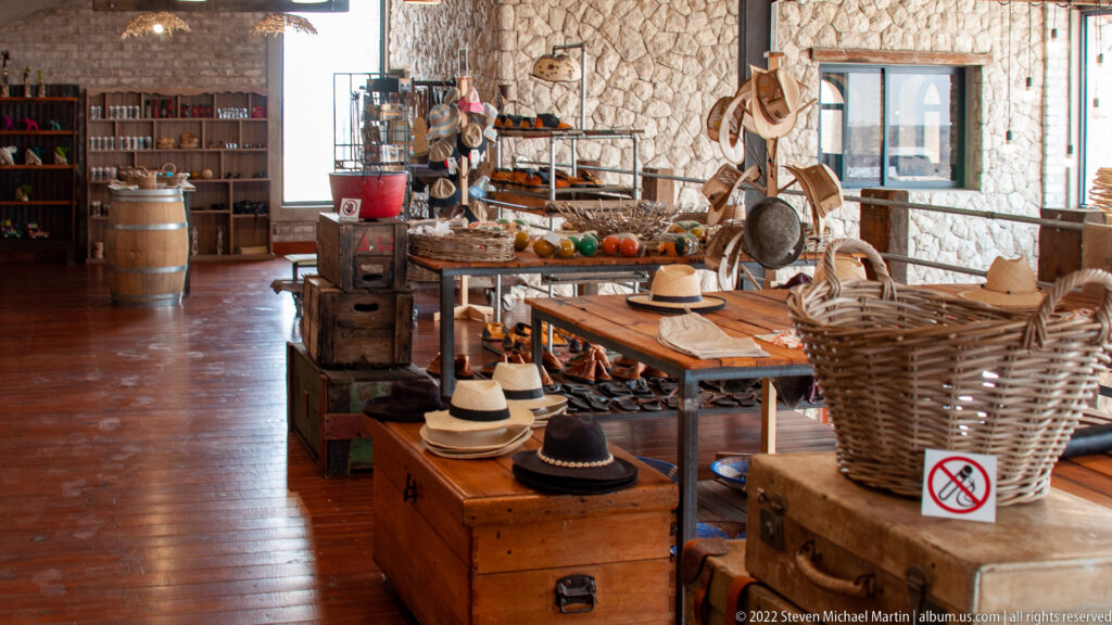 Rustic shop display with brimmed hats, woven baskets and accessories on wooden tables against pale stone walls.