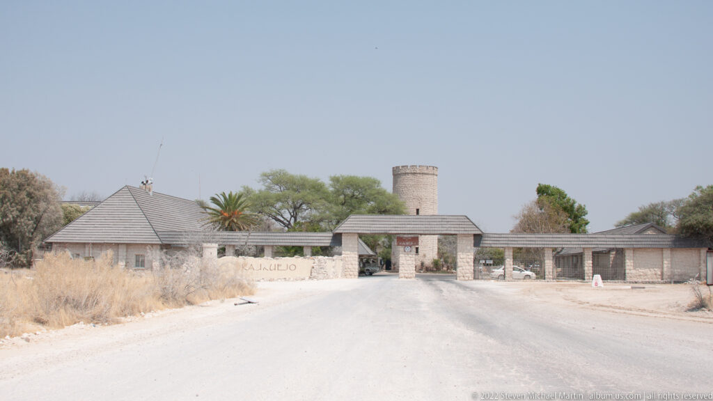 Road approaching the stone entrance buildings and cylindrical tower at Okaukuejo beneath a clear sky.
