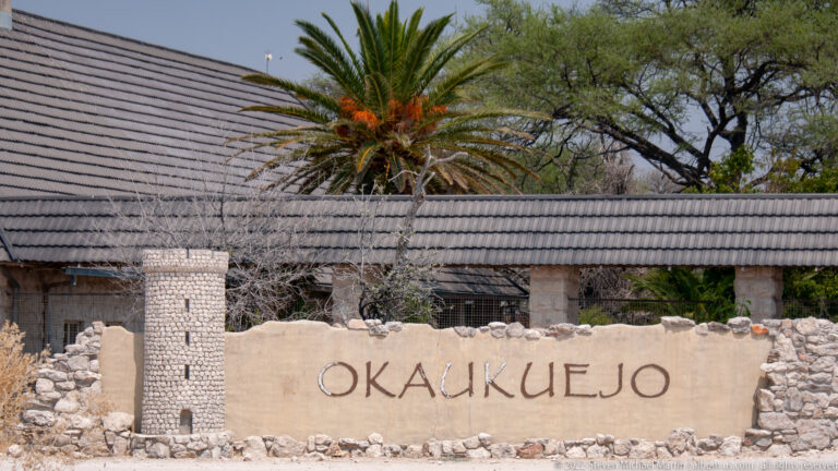 Stone entrance wall bearing the word OKAUKUEJO beside a small round tower and tiled-roof building.