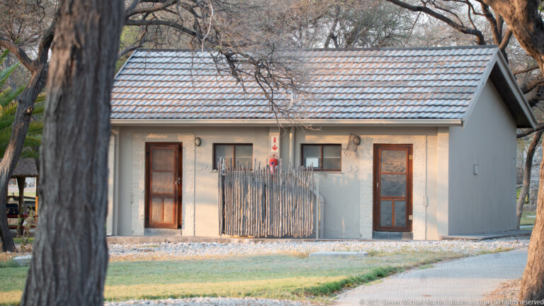 Small pale guest bungalow with tiled roof, two doors, trees, and a reed privacy screen at Okaukuejo.