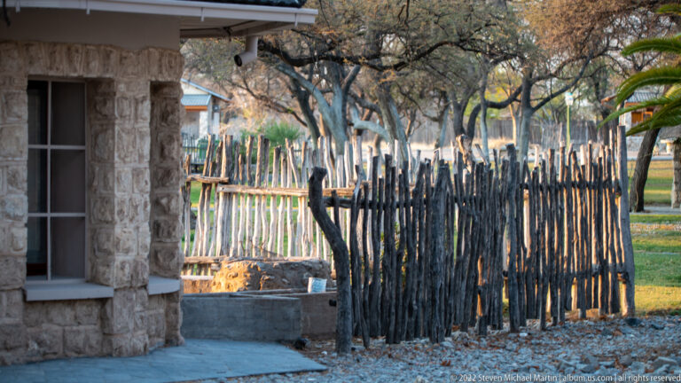 Corner of a stone building beside a tall irregular wooden-pole fence under trees at Okaukuejo.