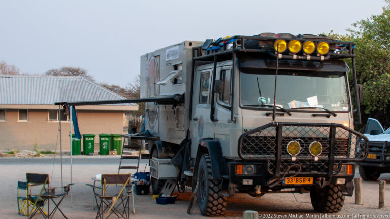 Large gray expedition truck with roof gear, auxiliary lights, protective grille, ladder, and folding chairs parked at camp.