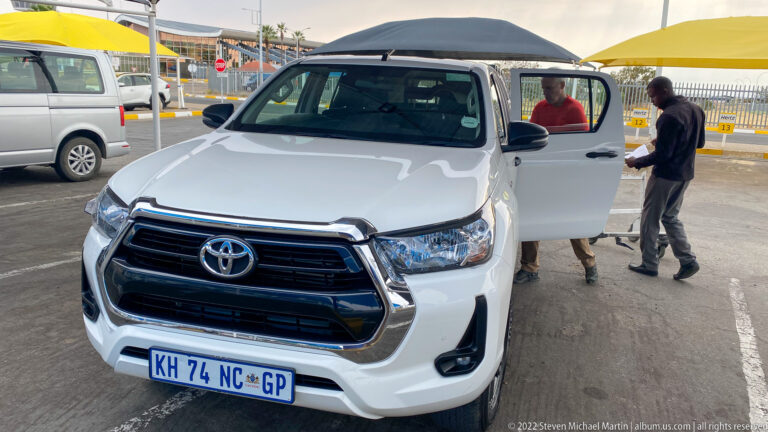 White Toyota Hilux pickup with passenger door open while two men stand beside it at a paved pickup area.