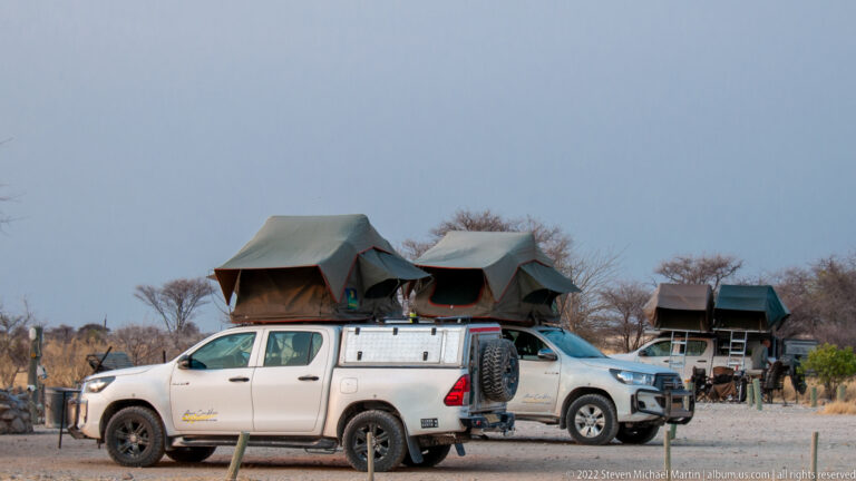 Two white pickup trucks fitted with raised canvas rooftop tents and safari cargo canopies in a dry campground.