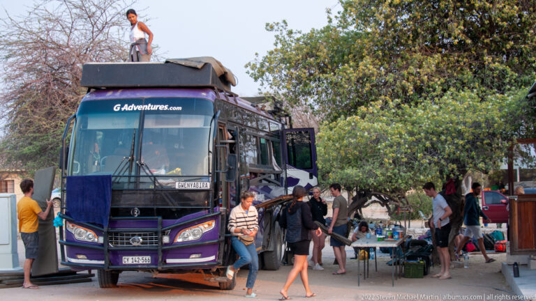 Dark G Adventures coach with open compartments, travelers, and luggage gathered beside it under trees.