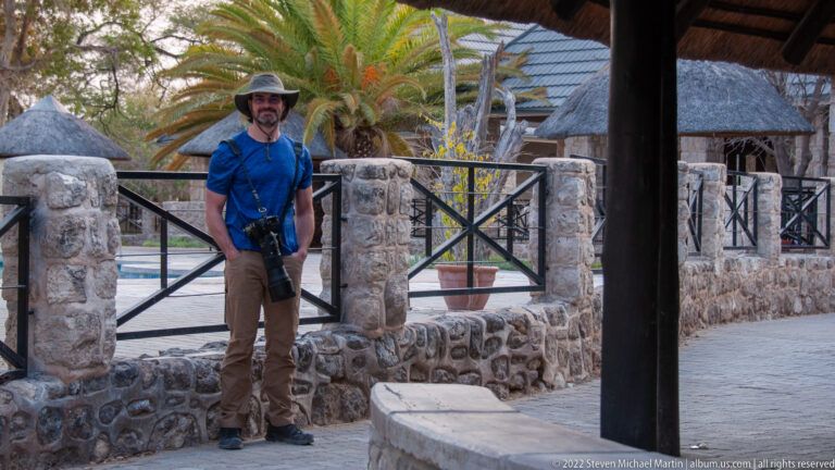 Man in safari clothing holding a long-lens camera beside stone walls, palms, and thatched lodge structures.