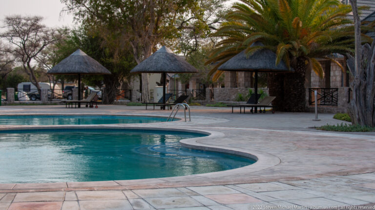 Curving turquoise swimming pool bordered by pale paving, lounge chairs, palm trees, and small thatched shelters.